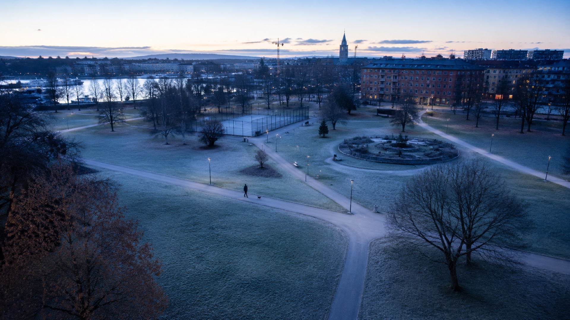 Kommunal park i Stockholm i gryningsljus, en person rastar hund längs gångstigen